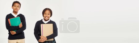 Photo for Happy african american schoolkids in neat uniform holding textbooks and standing on grey, banner - Royalty Free Image