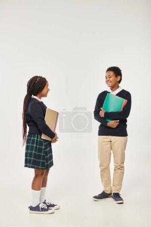 heureux afro-américains enfants en uniforme scolaire soigné tenant des manuels et debout sur fond gris