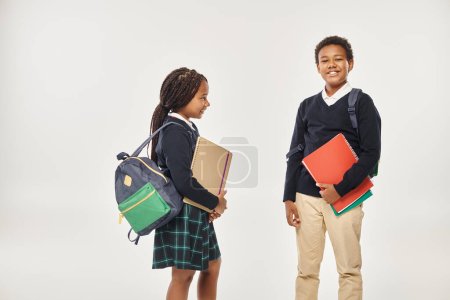 alegre afroamericano niños en uniforme escolar celebración de libros de texto y de pie sobre gris telón de fondo