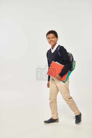 feliz afroamericano colegial en uniforme sosteniendo cuadernos y de pie sobre fondo gris