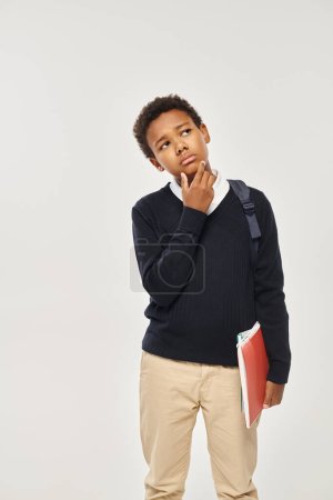 nachdenklicher afrikanisch-amerikanischer Schuljunge in Uniform mit Notizbüchern vor grauem Hintergrund