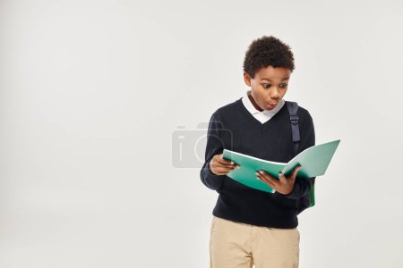 sorprendido colegial afroamericano en uniforme mirando notas en libro de texto sobre fondo gris