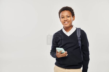 happy african american schoolboy in uniform holding smartphone and standing on grey background