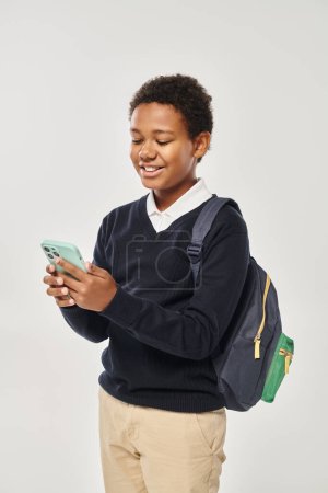 colegial afroamericano feliz en uniforme usando el teléfono inteligente y de pie sobre fondo gris