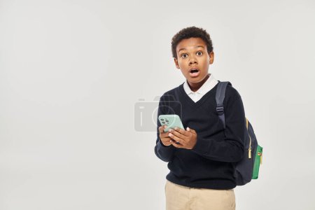 shocked african american schoolboy in uniform holding smartphone and standing on grey background