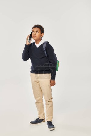 Photo for African american schoolboy in uniform talking on smartphone and standing on grey background - Royalty Free Image