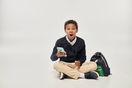 Schockierter afrikanisch-amerikanischer Junge in Uniform nutzt Smartphone und sitzt neben Rucksack vor grauem Hintergrund