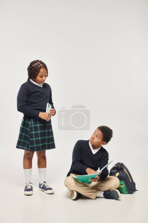 afrikanisch-amerikanische Schülerin hält Smartphone in der Nähe von Schüler beim Lernen mit Notizbuch auf grau