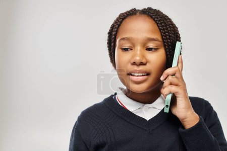 Photo for Preteen african american schoolgirl in uniform talking on smartphone on grey background, look down - Royalty Free Image