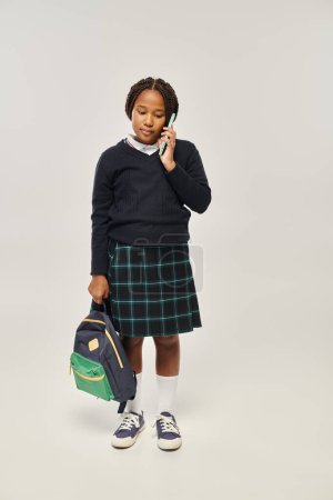 Photo for Preteen african american schoolgirl in uniform talking on smartphone and holding backpack on grey - Royalty Free Image