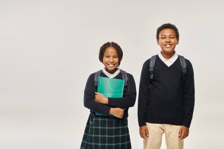 happy african american schoolgirl holding textbook near boy in sweater on grey background