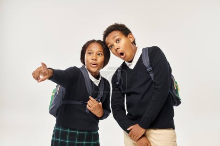 shocked african american schoolgirl poising away while standing near boy on grey background