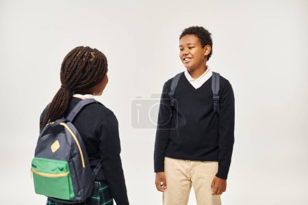 fröhliche afrikanisch-amerikanische Schulkinder mit Rucksäcken stehen und plaudern auf grauem Hintergrund