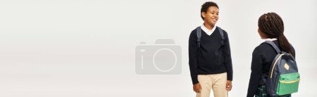 Photo for Cheerful african american schoolkids with backpacks standing and chatting on grey background, banner - Royalty Free Image