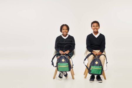 fröhliche afrikanisch-amerikanische Schulkinder mit Rucksäcken, die vor grauem Hintergrund sitzen und in die Kamera schauen