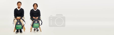Photo for Cheerful african american schoolkids with backpacks sitting and looking at camera on grey, banner - Royalty Free Image