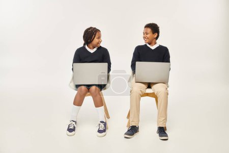 fröhliche afrikanisch-amerikanische Schulkinder in Uniform mit Laptops vor grauem Hintergrund