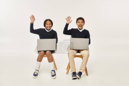 excited african american schoolkids in uniform using laptops and waving hands on grey backdrop