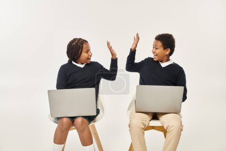 feliz afroamericano colegiales en uniforme usando computadoras portátiles y dando cinco alto en gris telón de fondo