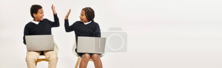 Photo for Happy african american schoolkids in uniform using laptops and giving high five on grey, banner - Royalty Free Image