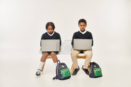 focused african american schoolkids in uniform using laptops and sitting on grey background