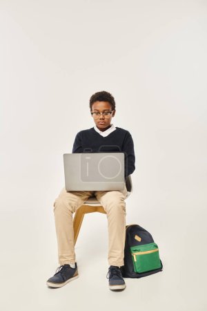 focused african american schoolboy in uniform and glasses using laptop and sitting on grey backdrop