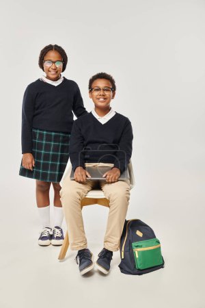 fröhliches afrikanisch-amerikanisches Mädchen in Uniform steht neben einem Schuljungen mit Laptop vor grauem Hintergrund