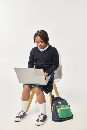 focalisé afro-américaine écolière en uniforme et des lunettes à l'aide d'un ordinateur portable et assis sur fond gris