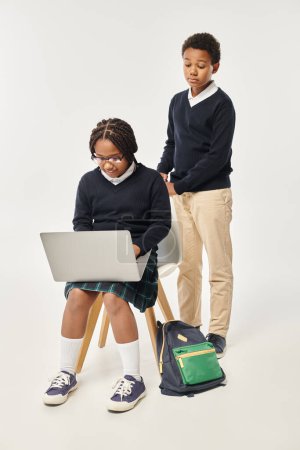 african american boy in uniform standing near excited schoolgirl using laptop on grey backdrop