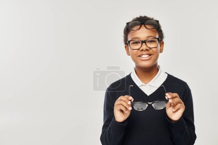 optimistic african american boy in sweater and eyewear holding glasses and looking at camera on grey tote bag #692618686