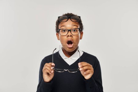 shocked african american boy in sweater and eyewear holding glasses and looking at camera on grey