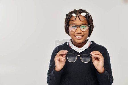 happy african american girl in sweater and eyewear holding glasses and looking at camera on grey