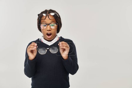 shocked african american girl in sweater and eyewear holding glasses and looking at camera on grey