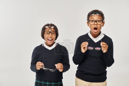 astonished and surprised african american schoolchildren in eyewear on grey background, reaction