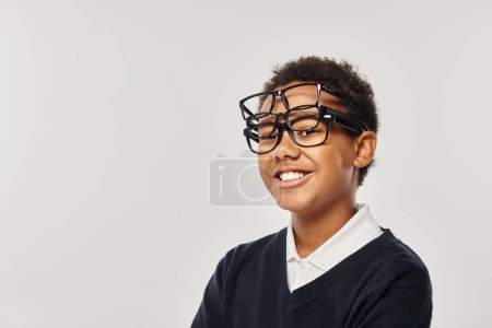 optimista afroamericano chico en suéter y gafas sosteniendo gafas y mirando a la cámara en gris