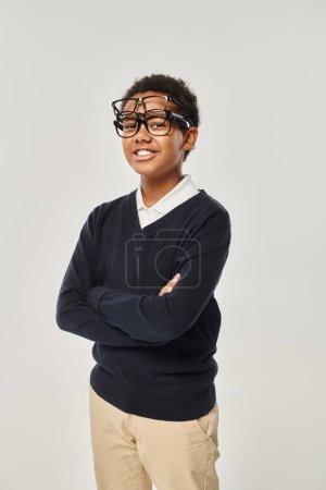 cheerful african american boy in sweater and eyewear holding glasses and looking at camera on grey