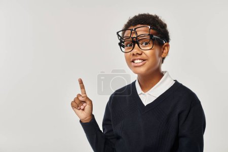 Téléchargez les photos : Écolier afro-américain optimiste en lunettes tenant des lunettes et regardant la caméra sur gris - en image libre de droit