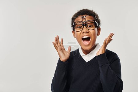 excited african american kid in sweater and eyewear holding glasses and looking at camera on grey