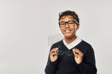 happy african american schoolboy in eyewear holding glasses and looking at camera on grey backdrop mug #692618876