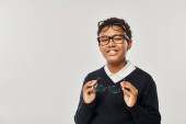 happy african american schoolboy in eyewear holding glasses and looking at camera on grey backdrop Sweatshirt #692618876