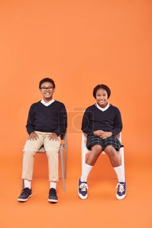 heureux afro-américains écoliers en uniforme souriant et assis sur des chaises sur fond orange