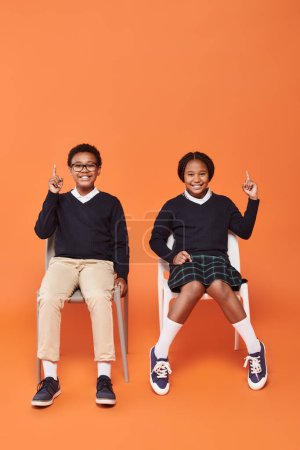 fröhliche afrikanisch-amerikanische Schulkinder in Uniform, die nach oben zeigen und auf Stühlen vor orangefarbenem Hintergrund sitzen