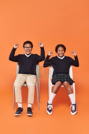 excités écoliers afro-américains en uniforme souriant et assis sur des chaises sur fond orange