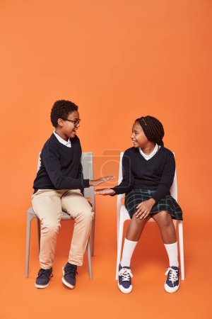 cheerful african american schoolkids in uniform sitting on chairs and giving five on orange backdrop