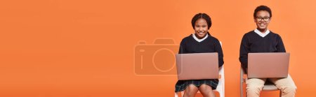 fröhliche afrikanisch-amerikanische Schulkinder in Uniform auf Stühlen sitzend und mit Laptops auf orangefarbenem Banner