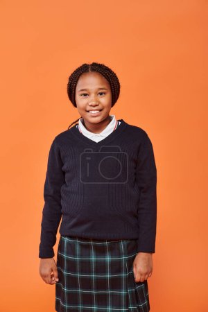 happy african american schoolgirl in uniform smiling and looking at camera on orange background