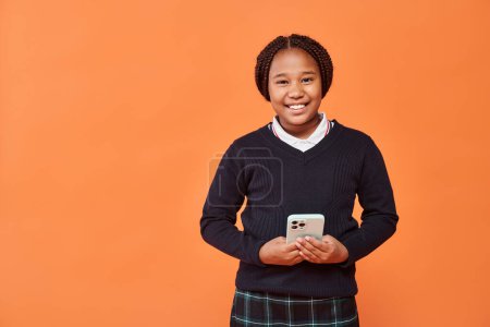 happy african american schoolgirl in uniform smiling and holding smartphone on orange background