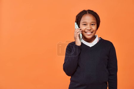 happy african american schoolgirl in uniform smiling and talking on smartphone on orange background