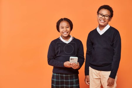 cheerful african american schoolkids in uniform looking at smartphone on orange background