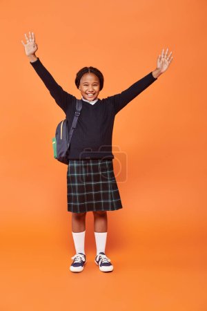 cheerful african american schoolgirl in uniform smiling and holding backpack on orange background tote bag #692619056